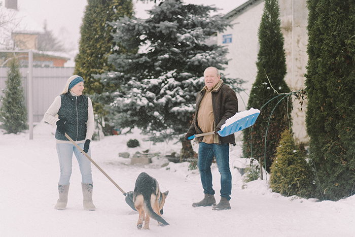 Image of couple shoveling snow with their dog. Consider hiring a snow removal professional